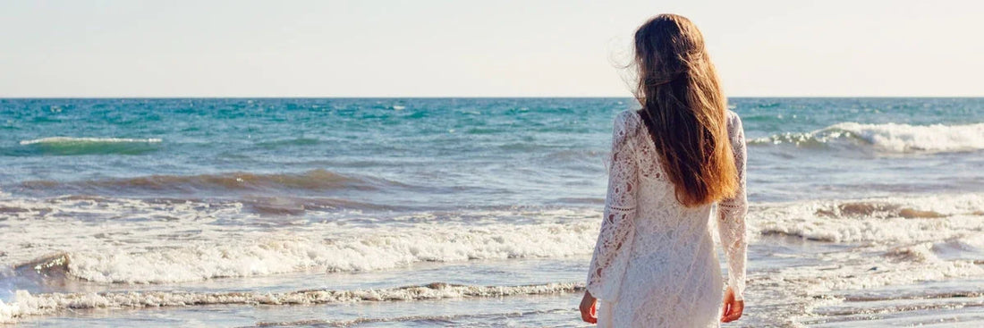 Woman in white lace dress walking on beach, ocean waves behind her