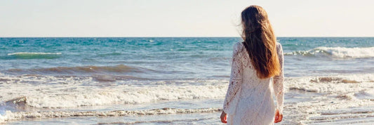 Woman in white lace dress walking on beach, ocean waves behind her