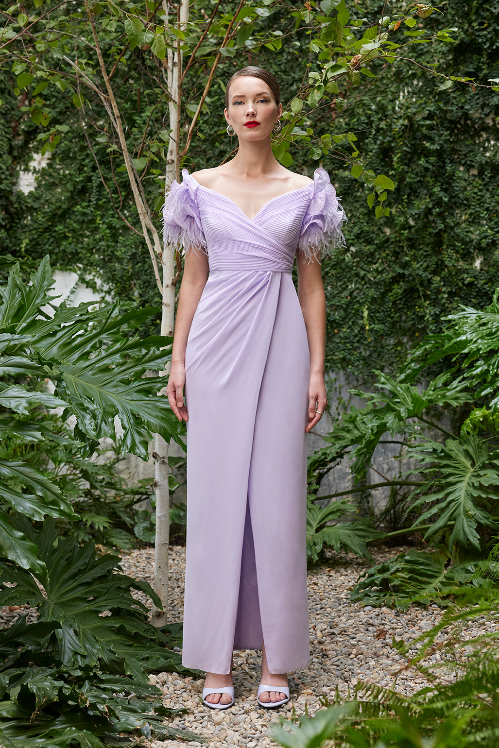 Model presenting a lilac evening gown (offered in dark fuchsia and turquoise as well) with feathered shoulders and a thigh-high slit, posing in a lush garden.