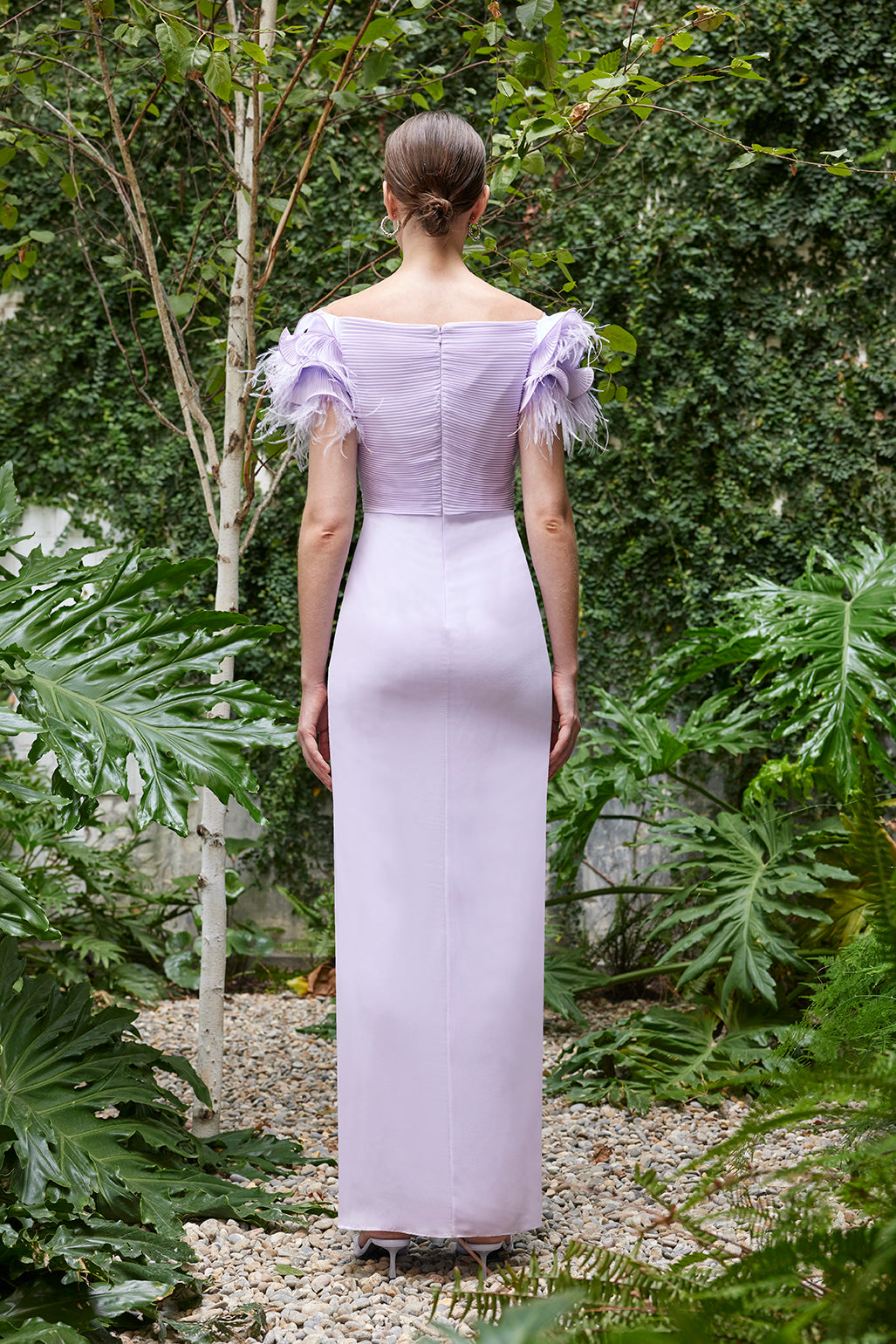 Back view of an elegant evening gown with feathered shoulders, available in lilac, dark fuchsia, and turquoise colors, showcased in a tranquil garden.