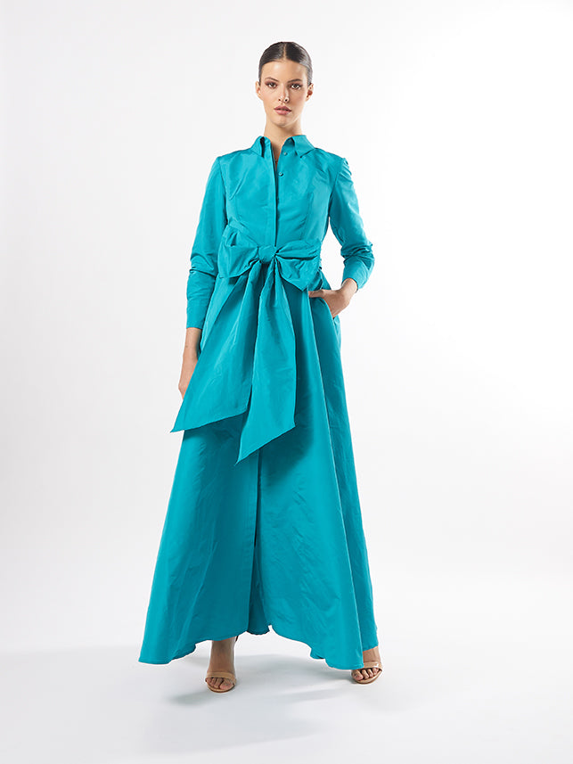 A model displays a teal silk wrap dress with a tie waist, also available in fuchsia and pistachio, standing against a plain white background.