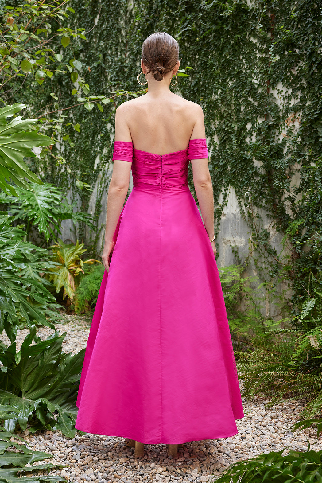 Back view of the fuchsia evening gown, also offered in red, showcasing the smooth zipper closure and off-the-shoulder sleeves against a garden wall draped with ivy.