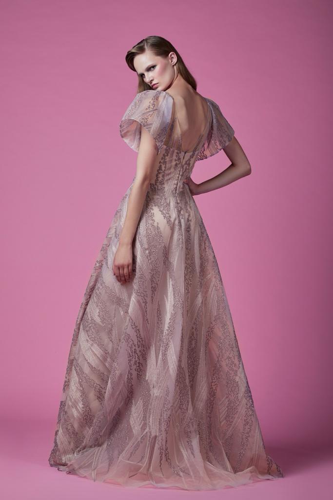 Rear view of a woman in a blush tulle gown with intricate embroidery and a graceful silhouette, against a pink backdrop, exuding vintage elegance.