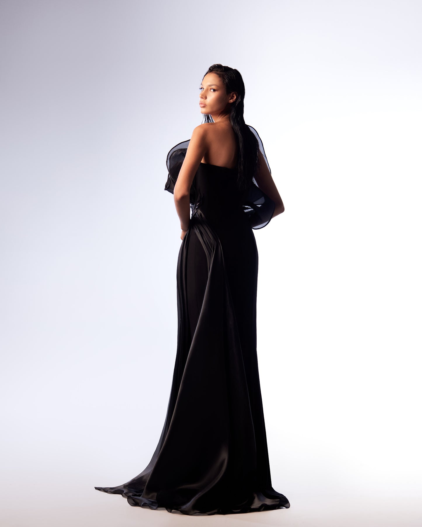 An elegant model displays the Enigmatic Noir Ruffle Gown, a black satin evening dress with a structured, sculptural ruffle bodice, and a high slit on the side, shown from multiple angles against a gradient background.